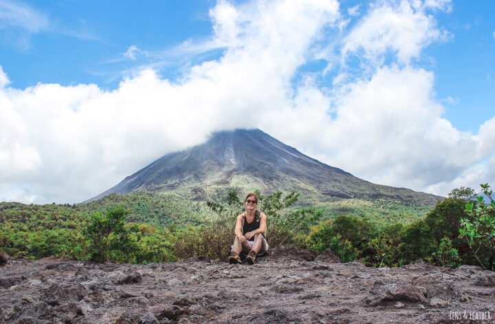 travel blogger Jessi sitting on cold lava trail in front of clouded volcano Arenal